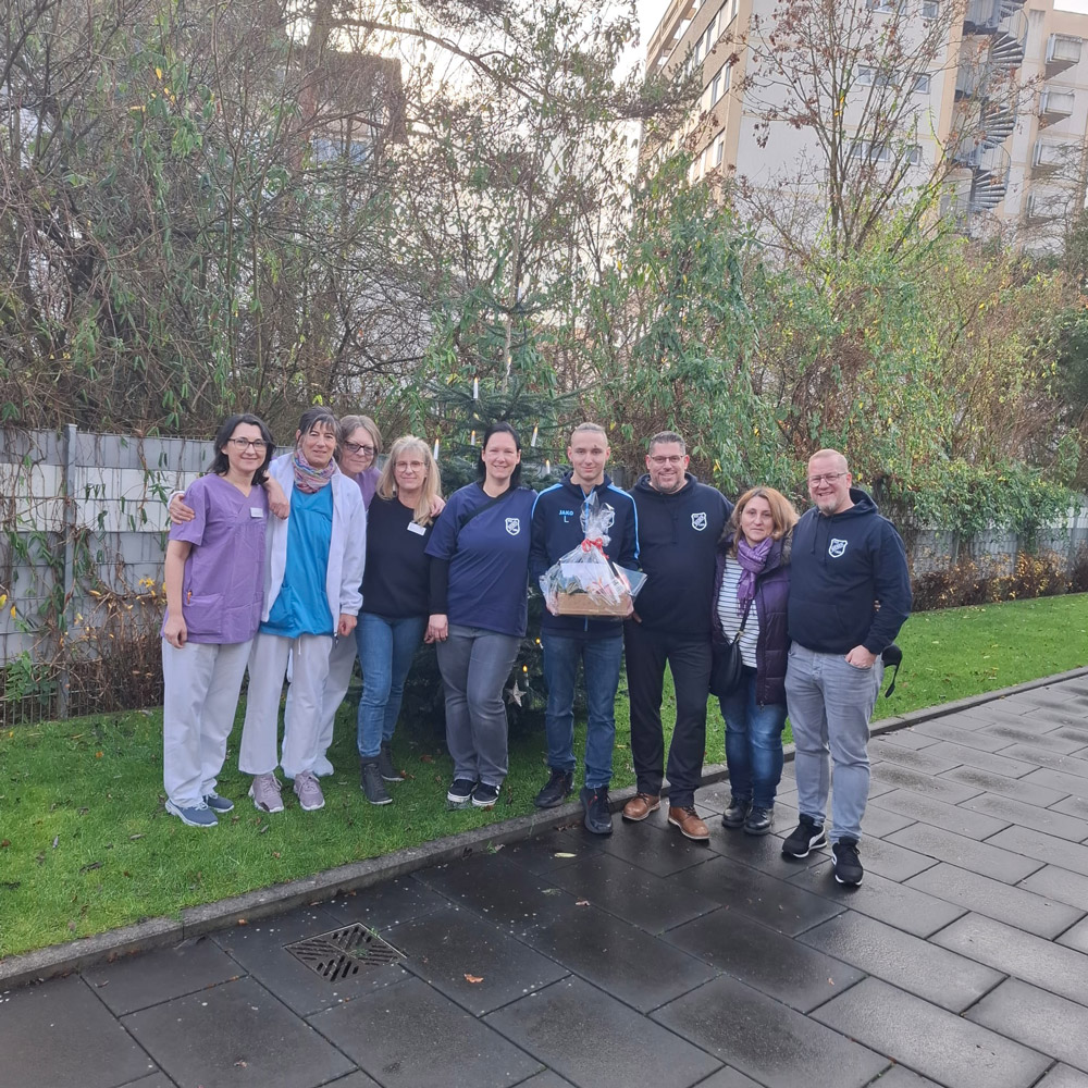 Eine Gruppe von acht Erwachsenen lächelt freundlich im Freien, eine Person hält einen Präsentkorb